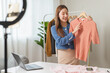 © Pormezz - social media market concept. Young Asian woman seller showing second-hand clothes during live streaming in the mobile application.