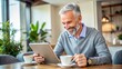 © Man888 - A smiling mature adult holds a digital tablet, making an online transaction, surrounded by coffee cups and a laptop, in a cozy morning atmosphere.