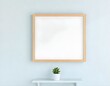 © Elie - A blank framed mockup hanging on a light blue pastel wall, with a small potted succulent on a shelf below create with ai