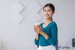 © David - Young successful employee business asian woman in blue shirt work hold takeaway delivery craft paper cup coffee to go stand at desk with laptop pc computer at office.