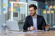 © Liubomir - Businessman in a suit working in modern office. Smiling while using a calculator and laptop, surrounded by sticky notes on glass wall. Represents accounting, finance, and business planning.
