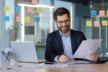 Wall Mural - Businessman with glasses and beard sits at office desk, reviewing documents with pen in hand. Surrounded by laptop, notes on window, displaying sense of productivity and focus.