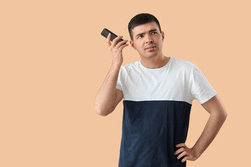 Wall Mural - Displeased young man with smartphone listening to voice message on beige background