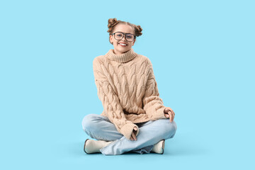 Sticker - Happy beautiful young woman in warm sweater and eyeglasses sitting on blue background