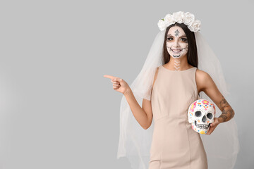 Poster - Young woman dressed as dead bride for Halloween party with painted human skull pointing at something on grey background