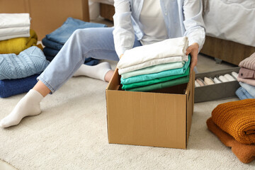 Young woman packing stack of clothes into cardboard box in bedroom, closeup