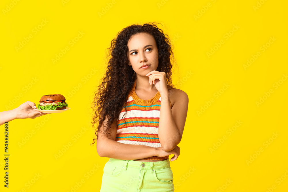 Thoughtful young African-American woman and hand with burger on yellow background. Diet concept