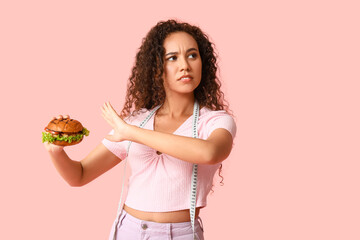 Wall Mural - Young African-American woman with measuring tape showing stop gesture against burger on pink background. Diet concept