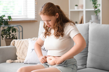 Young woman in tight shorts sitting on sofa at home. Weight gain concept