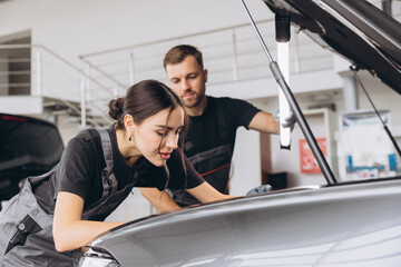 Trying to identify the source of the problem. Two mechanics man and woman working together at open hood on a car in an auto repair shop.