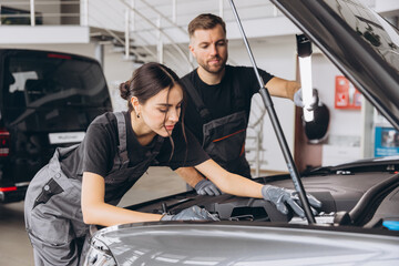 Trying to identify the source of the problem. Two mechanics man and woman working together at open hood on a car in an auto repair shop.