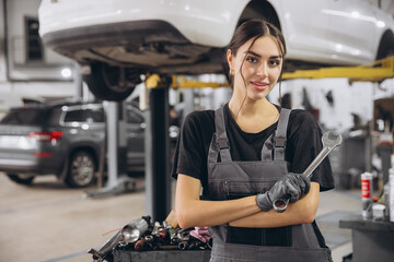 Wall Mural - Smiling young woman mechanic standing beneath lifted car, holding tools and looking at camera in car service garage