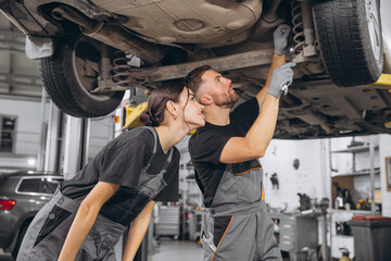 two focused professional mechanics, bearded man and young woman, wearing grey overalls, repairing un