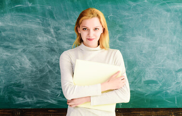 Wall Mural - Blonde student girl with book near blackboard in university. Smiling female teacher standing with teacher's journal in front of chalkboard. School teacher or lecturer with class journal in classroom.