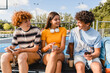 © InsideCreativeHouse - Group of teenagers students high school pupils classmates using gadgets holding smart mobile phones talking communicating hanging out sitting outside sport court yard. Friendship concept