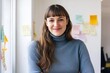 © 为轩 张 - American Woman with Long Brown Hair and Bangs in Blue Turtleneck Sweater Smiling at Camera, Standing in Office Space with Post-it Notes on White Wall and Natural Light from Window