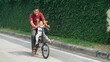 © indraprakkk - A father and son joyfully riding a bicycle along an ivy-covered wall in a quiet urban area. The warmth of family in the city