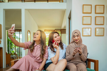 Three Indonesian women are sitting on a couch and two of them video call on their phones