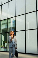 Wall Mural - Business woman strolls confidently outdoors in smart attire, checking her messages against the backdrop of a modern glass building on a sunny day