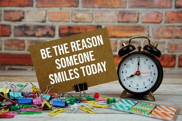 Wall Mural - be the reason someone smiles today text on paper card with alarm clock and stationary on wooden back
