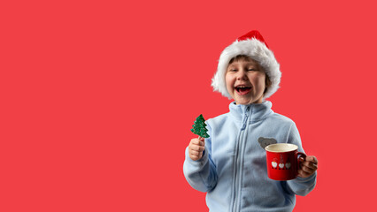 Laughing child in Santa Claus hat holding a Christmas tree shaped lollipop on a stick in one hand and a red cup in the other hand. The background is red. Copy space.