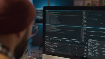 Over the shoulder view of male system administrator wearing beanie sitting at workplace coding, maintaining servers and software applications