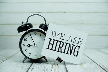 We Are Hiring text on paper card with alarm clock on wooden background