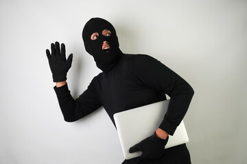 Wall Mural - Young Adult black masked robber is holding silver colored laptop. Running, fleeing and escaping pose. Concept for hacking, scamming, phishing.