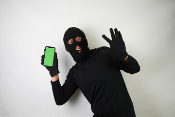 Wall Mural - Young Adult black masked robber is holding smartphone with blank green screen. Concept for hacking, scamming, phishing.