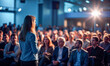 © vladnikon - A professional speaker stands confidently in front of a large audience, delivering an engaging presentation in a well-lit conference hall filled with attentive listeners.
