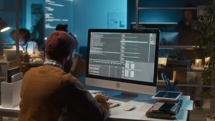 Timelapse back view of unrecognizable male software engineer wearing beanie sitting at computer writing new feature for web application. Workflow of team of IT specialists in background