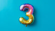 © KotBaton - Rainbow foil balloon number three. Top view. Colored number on blue background.