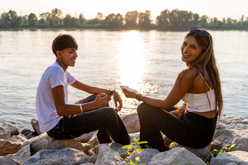 happy young teenage couple having fun using mobile phone outdoors sitting on white stones of a river bank