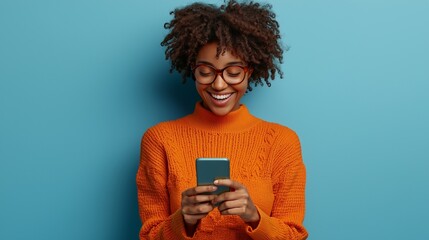 Photo of cheerful delighted african american woman types sms on modern cell phone device enjoys good internet connection dressed in orange jumper focused aside isolated on blue studio wall.