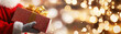 © PoodPhoto - Close-up of Santa Claus holding a wrapped Christmas gift with a golden bow, festive holiday lights in the background.