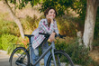 © ViDi Studio - Young happy minded woman she wearing checkered shirt casual clothes look camera walking stand lean on bicycle rest relax in spring sunshine green city park outdoors on nature. Urban leisure concept.