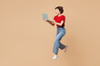 © ViDi Studio - Full body young smiling happy IT woman wear red t-shirt casual clothes jump high hold use work on laptop pc computer isolated on plain pastel light beige background studio portrait. Lifestyle concept.