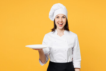 Wall Mural - Young surprised shocked excited chef cook baker woman wear white shirt black apron uniform toque chefs hat hold in hand empty plate isolated on plain yellow background studio. Cooking food concept.