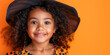 © julijadmi - A smiling young girl with curly hair wearing a witch costume, posing against an orange background, capturing a joyful Halloween moment.