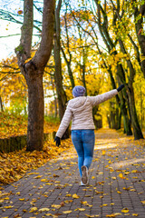 Wall Mural - Woman wearing jeans, beige jacket and winter hat walking on alley full of yellow fallen leaves in park on autumn day. Back view	