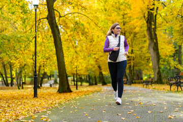 Wall Mural - Pretty middle-aged blonde woman dressed sporty running on alley full of beautiful autumnal trees in city park on autumn day. Front view. Autumnal training. Healthy running.