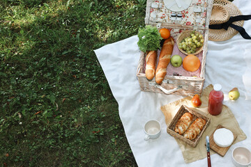 Poster - Picnic wicker baskets with delicious food, tableware and drink on white blanket outdoors, above view. Space for text