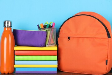 Backpack with different school stationery and bottle on wooden table against light blue background