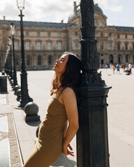 Young smiling attractive slim long haired female tourist walking in Paris in Louvre in summer sunny day, enjoying beautiful architecture and atmosphere of ancient city