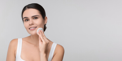 Poster - Young woman cleaning her face with cotton pad on light grey background. Banner design with space for text