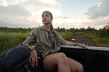 Young serene woman in casual attire looking at surrounding nature while sitting in body of pickup truck and enjoying summer trip