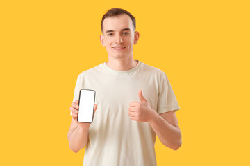 Wall Mural - Young man with mobile phone showing thumb-up on yellow background