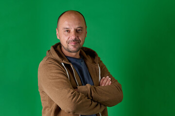 middle aged bald man in casual attire with crossed arms poses confidently against a green studio bac