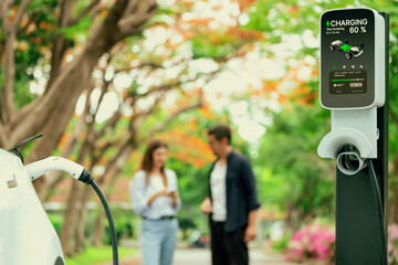 Focused EV car recharging battery on blurred background of lovey couple during autumnal road trip travel with electric vehicle recharging battery. Eco friendly travel on vacation during autumn. Exalt