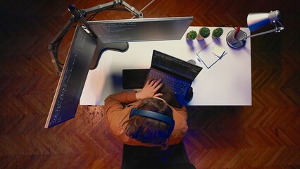 Top down view of engineer typing code in home office, using Java programming language to develop software application. It specialist using multimonitor PC setup and laptop to finish tasks, aerial shot